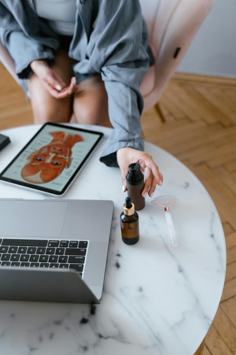 Top-down view of skincare products and jade roller on a table near a laptop and a tablet showing skincare routine.